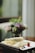 Delicate crepes are neatly arranged on a white plate, garnished with a small, vibrant parfait and colorful flowers. A blurred background shows soft light filtering through a window, with subtle hints of green and purple from out-of-focus plants or flowers.