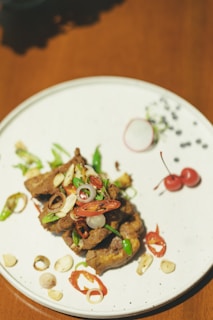 A plate presenting a dish comprised of seasoned meat garnished with sliced red and green chili peppers, scallions, and garlic slices. The arrangement includes a pair of bright red cherries on the side and a radish garnish, with the dish resting on a white plate set on a wooden table.