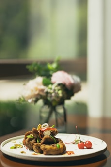 A beautifully arranged dish of crispy fried food garnished with fresh vegetables and herbs on a white plate. The setting includes a soft-focus background with a vase of flowers on a wooden table, creating a sophisticated and elegant dining atmosphere.
