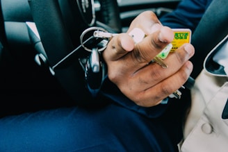 A close-up of a hand holding a set of keys with various keychains inside a vehicle. The steering wheel and part of a person's attire are visible, suggesting they might be in the driver's seat.