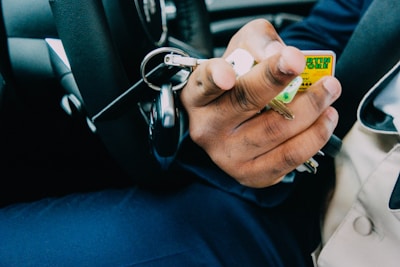 A close-up of a hand holding a set of keys with various keychains inside a vehicle. The steering wheel and part of a person's attire are visible, suggesting they might be in the driver's seat.