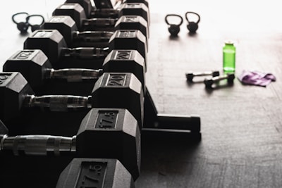 A row of hexagonal dumbbells is neatly arranged on a rack in the foreground, with numbers indicating their weights. In the background, there are kettlebells, a green water bottle, a pair of smaller dumbbells, and a purple towel on the floor, all set against a gym floor setting.