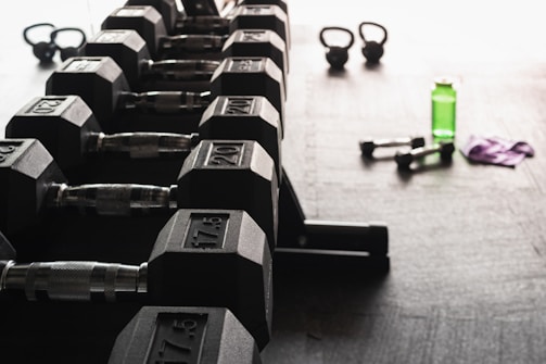 A row of hexagonal dumbbells is neatly arranged on a rack in the foreground, with numbers indicating their weights. In the background, there are kettlebells, a green water bottle, a pair of smaller dumbbells, and a purple towel on the floor, all set against a gym floor setting.