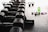 A row of hexagonal dumbbells is neatly arranged on a rack in the foreground, with numbers indicating their weights. In the background, there are kettlebells, a green water bottle, a pair of smaller dumbbells, and a purple towel on the floor, all set against a gym floor setting.
