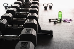 A row of hexagonal dumbbells is neatly arranged on a rack in the foreground, with numbers indicating their weights. In the background, there are kettlebells, a green water bottle, a pair of smaller dumbbells, and a purple towel on the floor, all set against a gym floor setting.