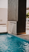 A modern water feature pours a small cascade into a swimming pool. The structure is sleek, made from a dark material, contrasting with the light blue water. The surrounding area appears to be a clean, minimalistic patio with bright lighting and a few potted plants in the background.
