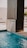 A modern water feature pours a small cascade into a swimming pool. The structure is sleek, made from a dark material, contrasting with the light blue water. The surrounding area appears to be a clean, minimalistic patio with bright lighting and a few potted plants in the background.