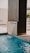 A modern water feature pours a small cascade into a swimming pool. The structure is sleek, made from a dark material, contrasting with the light blue water. The surrounding area appears to be a clean, minimalistic patio with bright lighting and a few potted plants in the background.