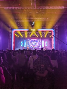A vibrant concert scene with a large crowd in front of a stage. Bright yellow spotlights shine down onto the stage, which features a colorful, circular LED backdrop. The atmosphere is filled with excitement and energy as people enjoy the live performance.