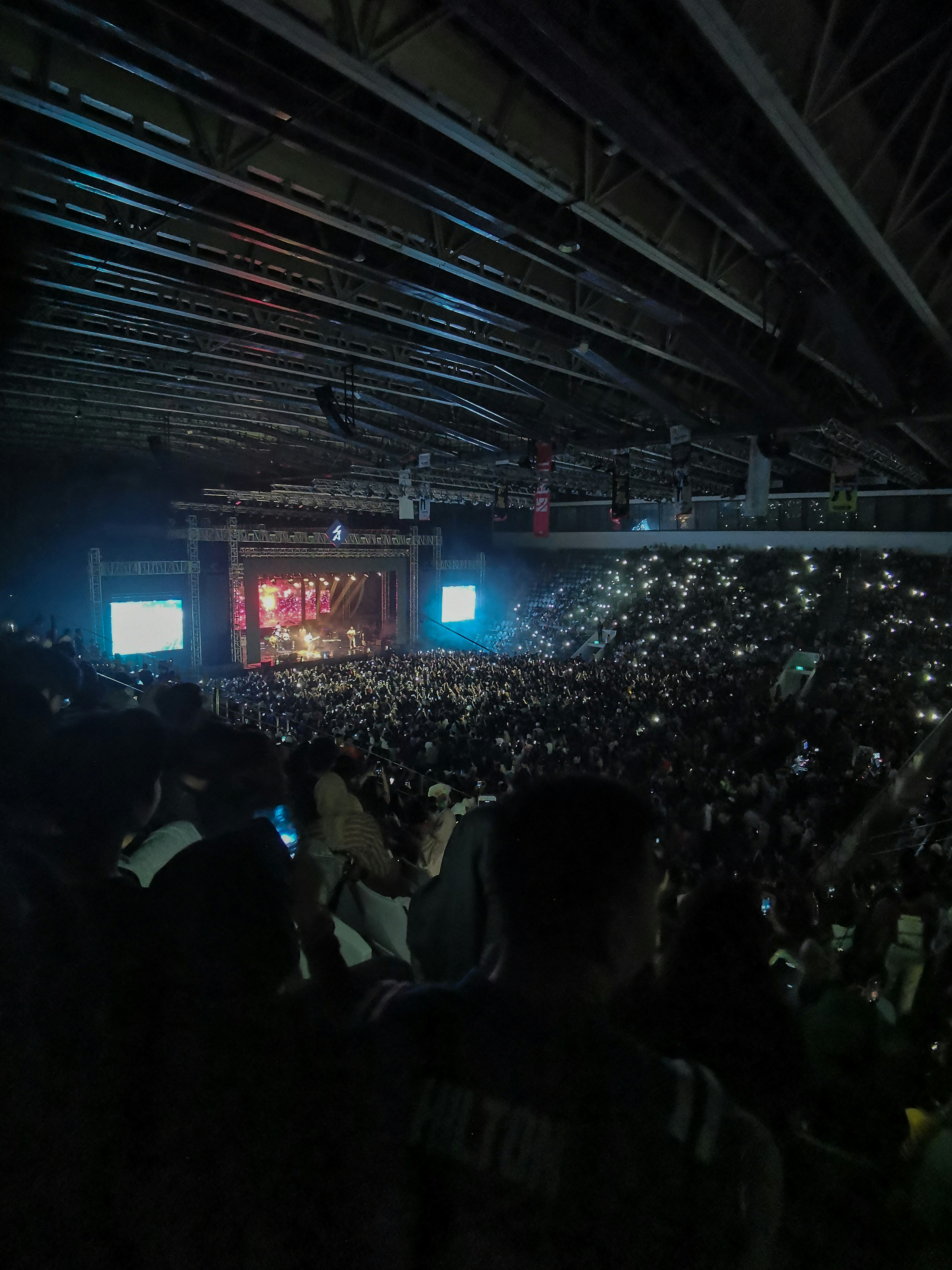 Massive concert crowd with colorful stage lighting, energetic atmosphere, thousands of fans with phone lights