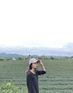 A person wearing a cap is standing in front of a vast tea plantation with rows of green plants stretching into the distance. The backdrop features rolling hills and a cloudy sky, suggesting a serene rural environment.