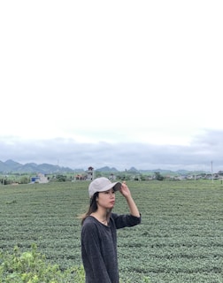 A person wearing a cap is standing in front of a vast tea plantation with rows of green plants stretching into the distance. The backdrop features rolling hills and a cloudy sky, suggesting a serene rural environment.