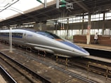 A sleek, modern bullet train stationed at a railway platform. The train is silver with blue accents, featuring the label 'JR 500 West Japan' on its side. Overhead wires and structures are visible, and the platform is equipped with safety rails and signage. A person in uniform is walking along the platform.