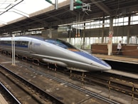A sleek, modern bullet train stationed at a railway platform. The train is silver with blue accents, featuring the label 'JR 500 West Japan' on its side. Overhead wires and structures are visible, and the platform is equipped with safety rails and signage. A person in uniform is walking along the platform.