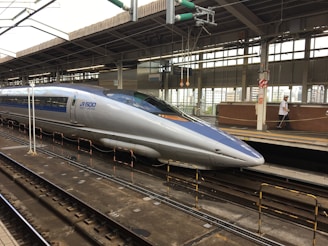 A sleek, modern bullet train stationed at a railway platform. The train is silver with blue accents, featuring the label 'JR 500 West Japan' on its side. Overhead wires and structures are visible, and the platform is equipped with safety rails and signage. A person in uniform is walking along the platform.
