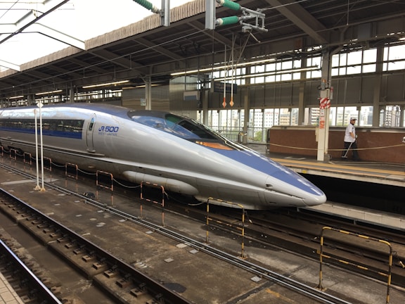 A sleek, modern bullet train stationed at a railway platform. The train is silver with blue accents, featuring the label 'JR 500 West Japan' on its side. Overhead wires and structures are visible, and the platform is equipped with safety rails and signage. A person in uniform is walking along the platform.