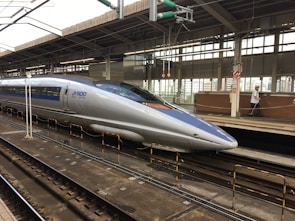 A sleek, modern bullet train stationed at a railway platform. The train is silver with blue accents, featuring the label 'JR 500 West Japan' on its side. Overhead wires and structures are visible, and the platform is equipped with safety rails and signage. A person in uniform is walking along the platform.