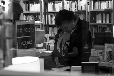 A person wearing a scarf and coat is browsing books in a store with shelves filled with books in the background. There is a sign labeled 'POLICIERS POLARS' and several books displayed on a table. The setting appears cozy and intimate, with subdued lighting.