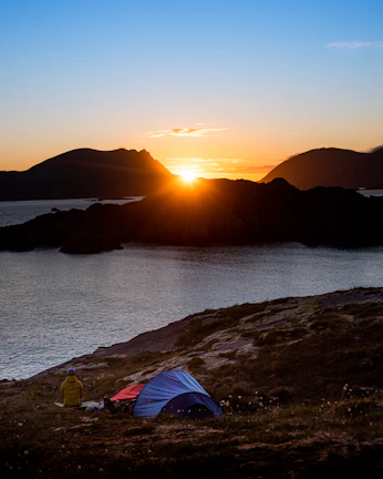 A serene tent site nestled by the water's edge with a vibrant orange sunset.