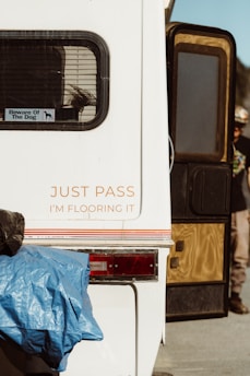 A white vehicle, likely a recreational vehicle or trailer, features a humorous sign with 'Just Pass, I'm Flooring It' printed in orange on its back. A window with blinds and a 'Beware of the Dog' sticker is visible. There is a blue tarp partially covering the rear, and part of a red taillight is noticeable. The side door is open, revealing a person partially in view wearing casual clothing.