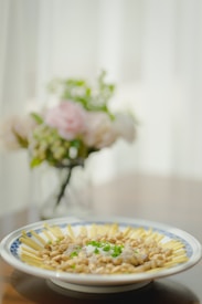 A decorative plate filled with a neatly arranged dish featuring various ingredients, including beans and sauces, garnished with chopped green onions. A blurred floral arrangement serves as the background, adding a touch of elegance.