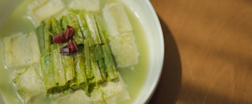 A dish featuring tofu pieces submerged in a light greenish broth. Strips of lettuce or cabbage are arranged neatly on top, garnished with small bits of red chili. The dish is served in a white bowl, placed on a wooden surface.