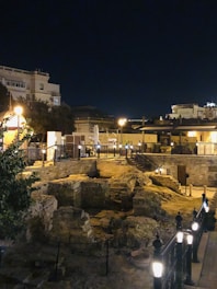 An illuminated archaeological site at night is surrounded by low stone walls and pathways, with nearby modern buildings casting a soft glow from streetlights. The area is fenced with decorative lights lining the perimeter.