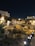 An illuminated archaeological site at night is surrounded by low stone walls and pathways, with nearby modern buildings casting a soft glow from streetlights. The area is fenced with decorative lights lining the perimeter.