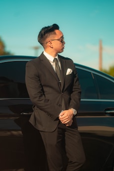 A person dressed in a formal suit leans against a dark-colored car under a clear blue sky, wearing sunglasses and a watch.