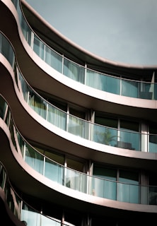 Curved balconies of a modern building with glass railings and reflections on the windows, showcasing architectural details and design. A potted plant is visible through the glass.