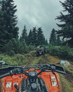 A vibrant orange ATV is pictured from a first-person view, traveling through a forest trail surrounded by dense green foliage and tall fir trees. The sky is overcast, contributing to a moody atmosphere as other ATVs can be seen ahead on the muddy path.