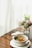 A white bowl filled with a variety of vegetables and tofu on a wooden table, next to a vase containing a bouquet of flowers. The scene appears to be by a window with sheer white curtains, allowing natural light to softly illuminate the space.
