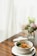 A white bowl filled with a variety of vegetables and tofu on a wooden table, next to a vase containing a bouquet of flowers. The scene appears to be by a window with sheer white curtains, allowing natural light to softly illuminate the space.