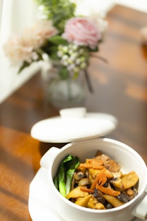 A dish in a white ceramic bowl featuring a medley of stir-fried vegetables, including mushrooms, carrots, and leafy greens. The bowl is placed on a wooden table with sunlight streaming in, creating a warm ambiance. In the background, there's a vase with a floral arrangement consisting of pink and white flowers with some greenery.