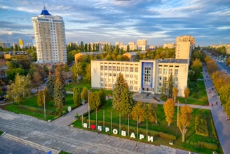 Aerial view of a cityscape with a prominent building in the center surrounded by trees and greenery. The building has a modern architectural style with a blue accent on the facade. In front of the building, there is a large sign with Cyrillic text and a heart symbol. Apartment buildings and trees are visible in the background under a partly cloudy sky.