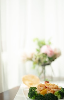 A plate of food featuring broccoli, shrimp, and possibly other vegetables sits on a table with a blurred background. The setting appears to be bright and airy, and there is a hint of floral arrangement in the background.