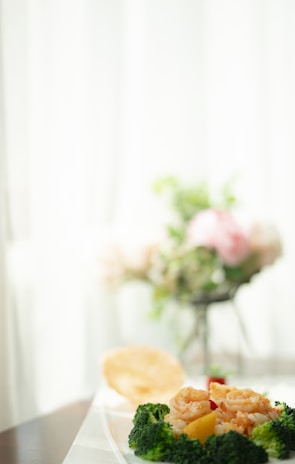 A plate of food featuring broccoli, shrimp, and possibly other vegetables sits on a table with a blurred background. The setting appears to be bright and airy, and there is a hint of floral arrangement in the background.