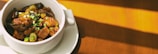 A white ceramic bowl filled with a savory dish sits on a matching small plate. The dish appears to contain pieces of meat, mushrooms, and is topped with chopped green onions. Sunlight casts warm shadows across a wooden surface in the background, highlighting the rich colors of the ingredients.