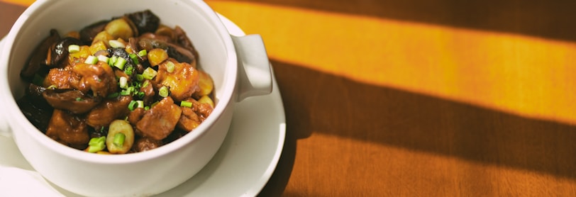 A white ceramic bowl filled with a savory dish sits on a matching small plate. The dish appears to contain pieces of meat, mushrooms, and is topped with chopped green onions. Sunlight casts warm shadows across a wooden surface in the background, highlighting the rich colors of the ingredients.