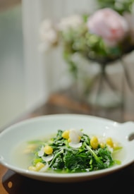A dish consisting of leafy green vegetables and yellow beans, garnished with white almond slices, served in a shallow white bowl. The background is softly blurred with hints of green and pink, suggesting a floral arrangement.