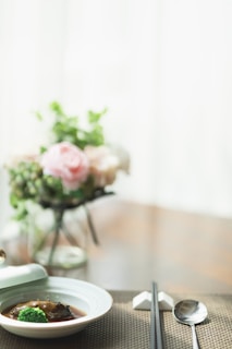 A minimalist table setting with a small dish containing a piece of meat and broccoli in sauce. In the background, there is a blurred vase with pastel-colored flowers, including a pink rose. The table is set with chopsticks and a spoon, resting on a woven placemat.