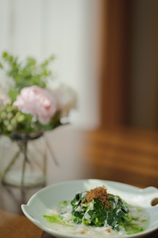 A beautifully arranged dish with a mound of green leafy vegetables drizzled with a creamy white sauce and topped with finely shredded brown garnish. It is presented on a white plate with a ruffled edge, sitting on a wooden table. In the soft-focus background, there's a vase containing pastel flowers, adding a touch of elegance and freshness.