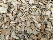 A closely packed arrangement of wood chips varying in size and shape, mostly in shades of brown and beige. Some chips have rough, textured surfaces while others appear smoother. There are a few small, scattered stones and a single, dried leaf among the chips.
