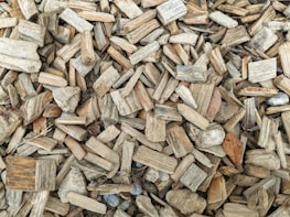A closely packed arrangement of wood chips varying in size and shape, mostly in shades of brown and beige. Some chips have rough, textured surfaces while others appear smoother. There are a few small, scattered stones and a single, dried leaf among the chips.