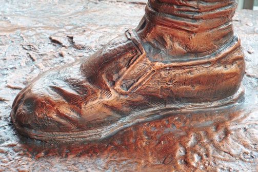 A bronze sculpture of a shoe, intricately detailed with creases and folds, showcasing craftsmanship. The surface appears textured and polished, with the shoe standing prominently on a similarly detailed base.