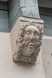 A detailed carving of a bearded face with curly hair and a mustache, integrated into an architectural structure. The stonework shows intricate detailing and craftsmanship.