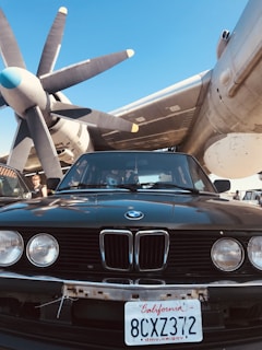 A vintage car with a California license plate is parked beneath the large propeller of an aircraft. The sky is clear and blue, creating a contrasting backdrop to the imposing machinery. A person is visible near the driver's side of the car.