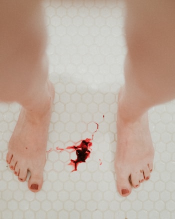 A pair of feet with painted toenails stands on a white hexagonal tiled floor, with red fluid resembling blood pooled on the surface.