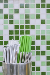 A metallic container holds green chopsticks, silver forks, and white napkins. The background is a tiled wall with varying shades of green and white tiles arranged in a square pattern.