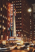 Bright neon lights illuminate a bustling city street. The iconic marquee of Radio City Music Hall glows in vibrant colors, while light trails from passing cars create a dynamic feel. Tall buildings surround the area, adding to the urban atmosphere.